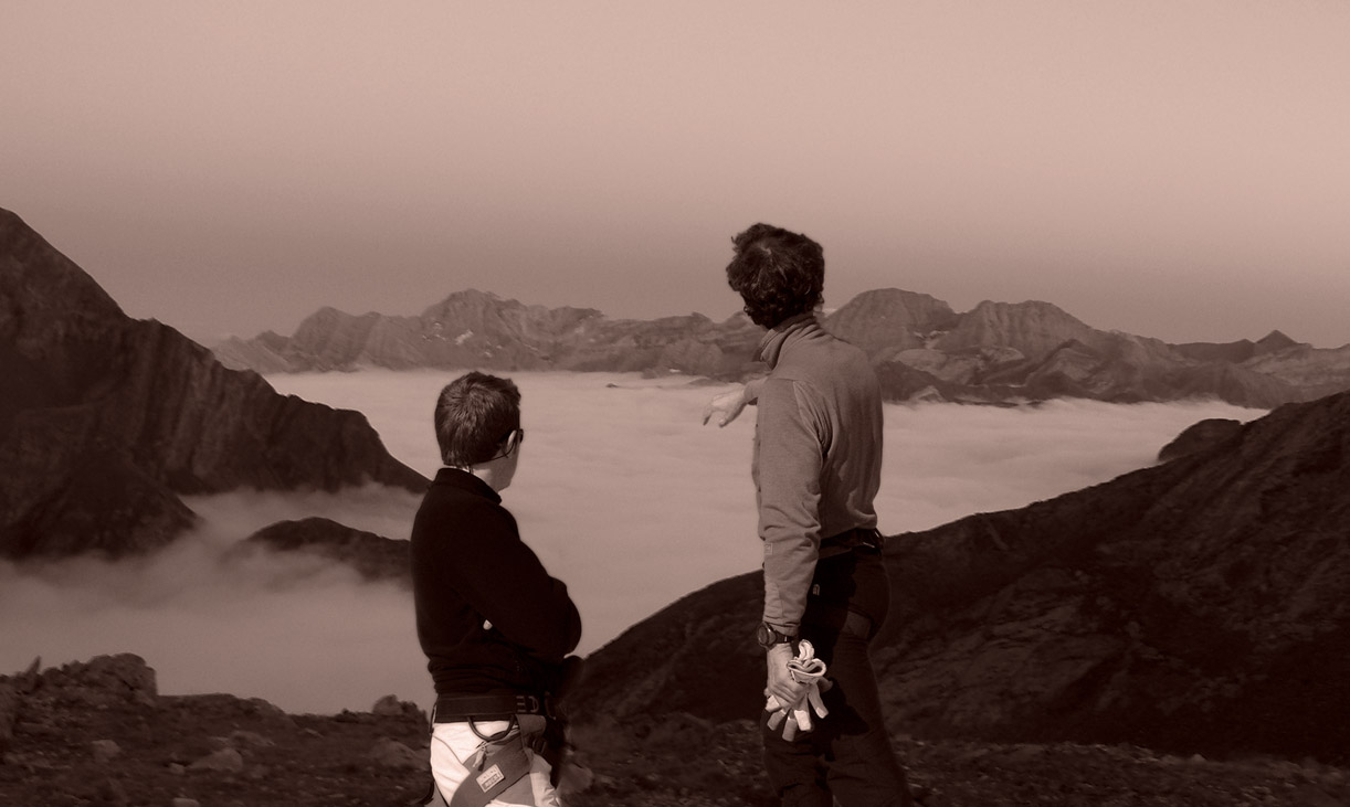 Two climbers on a summit above a sea of clouds, one pointing the way across